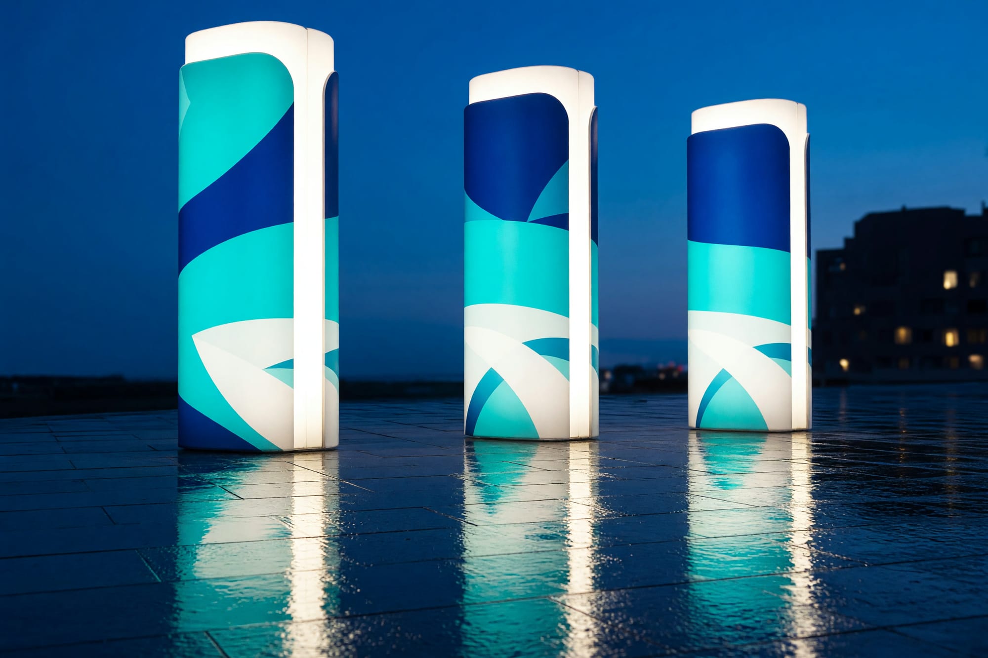 Aqua wayfinding totems on a wet plaza at dusk